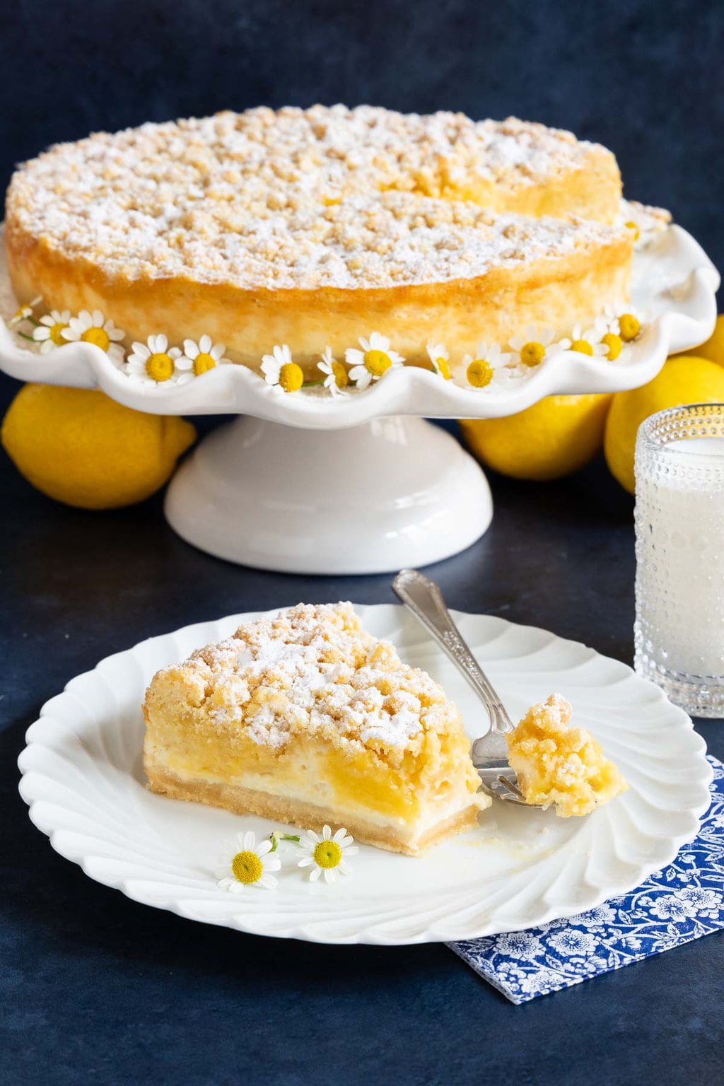 Vertical photo of a Italian Lemon Curd Ricotta Crumb Cake on a white pedestal stand with a slice of the cake on an individual serving plate in the foreground.