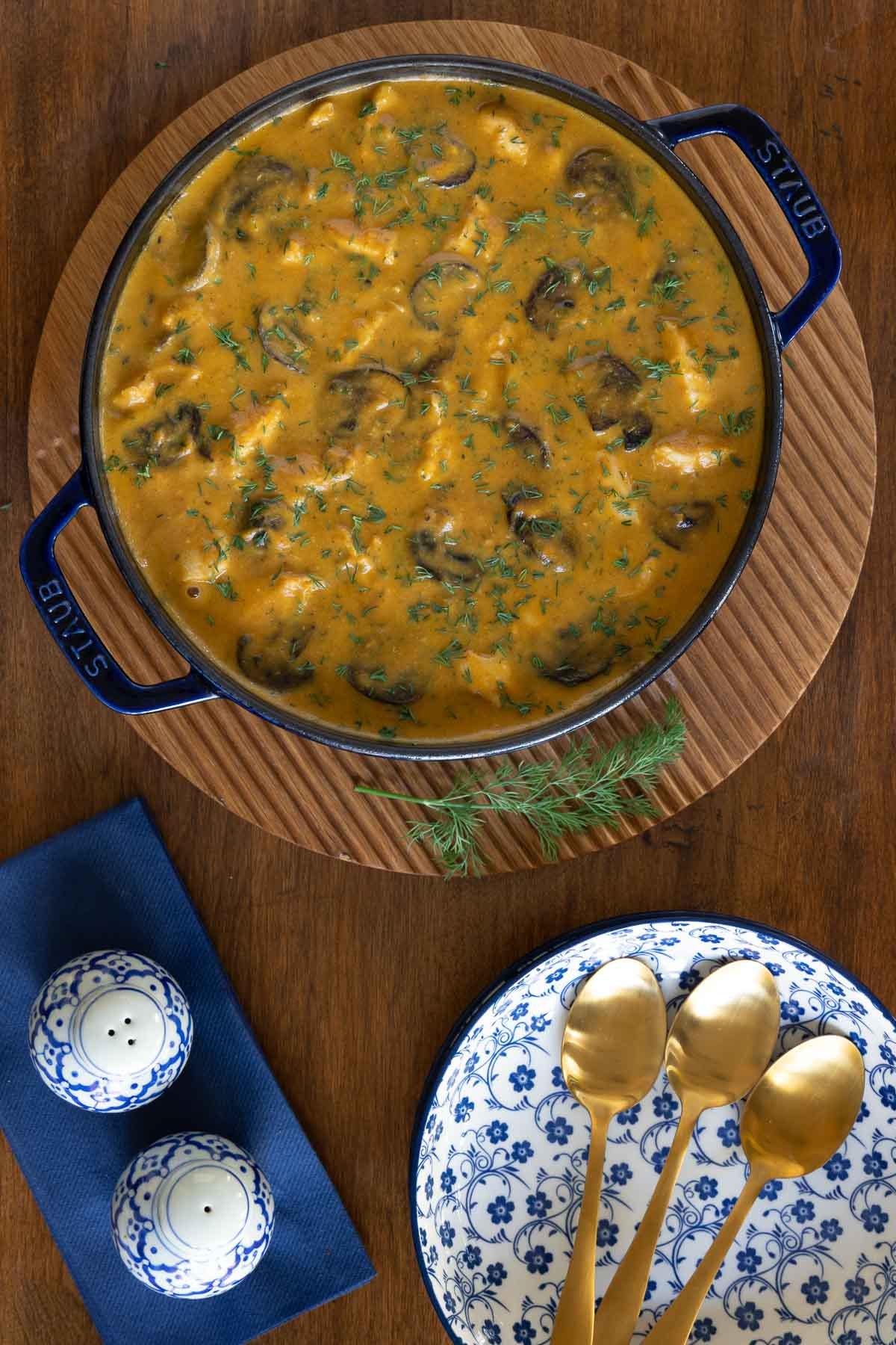 Vertical overhead photo of a blue cast iron pot of Low Carb Hungarian Mushroom Soup with chicken on a round wood serving board.