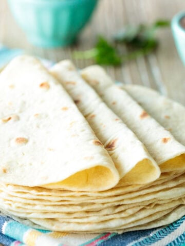 Horizontal closeup photo of a stack of Homemade Flour Tortillas on a colorful towel with a cup of salsa in the background.