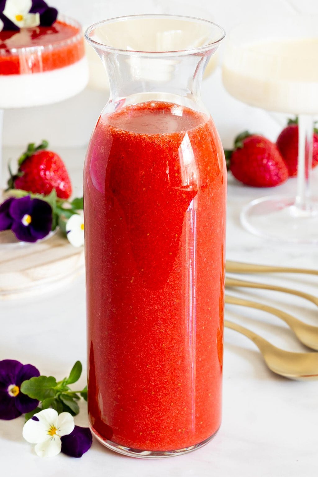Closeup of a jar of Strawberry Coulis with dessert glasses of Lemon Vanilla Bean Panna Cotta in the background along with fresh strawberries