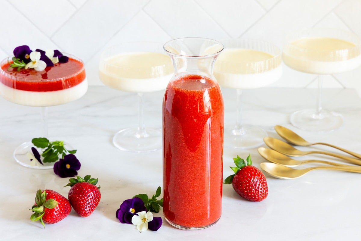 Horizontal photo of a jar of Ridiculously Easy Strawberry Coulis with glass dessert dishes of panna cotta in the background.