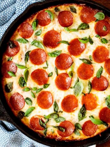 Horizontal overhead photo of a Deep Dish Pepperoni Pizza garnished with basil leaves on a wood table.