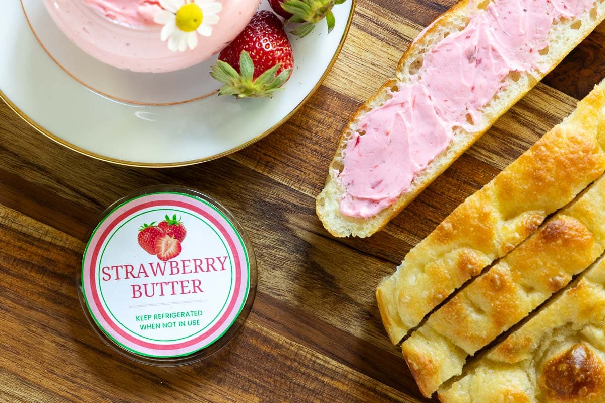Horzontal overhead closeup photo of a jar of Ridiculously Easy Fresh Strawberry Butter with a custom label for gift giving and a loaf of Focaccia bread.