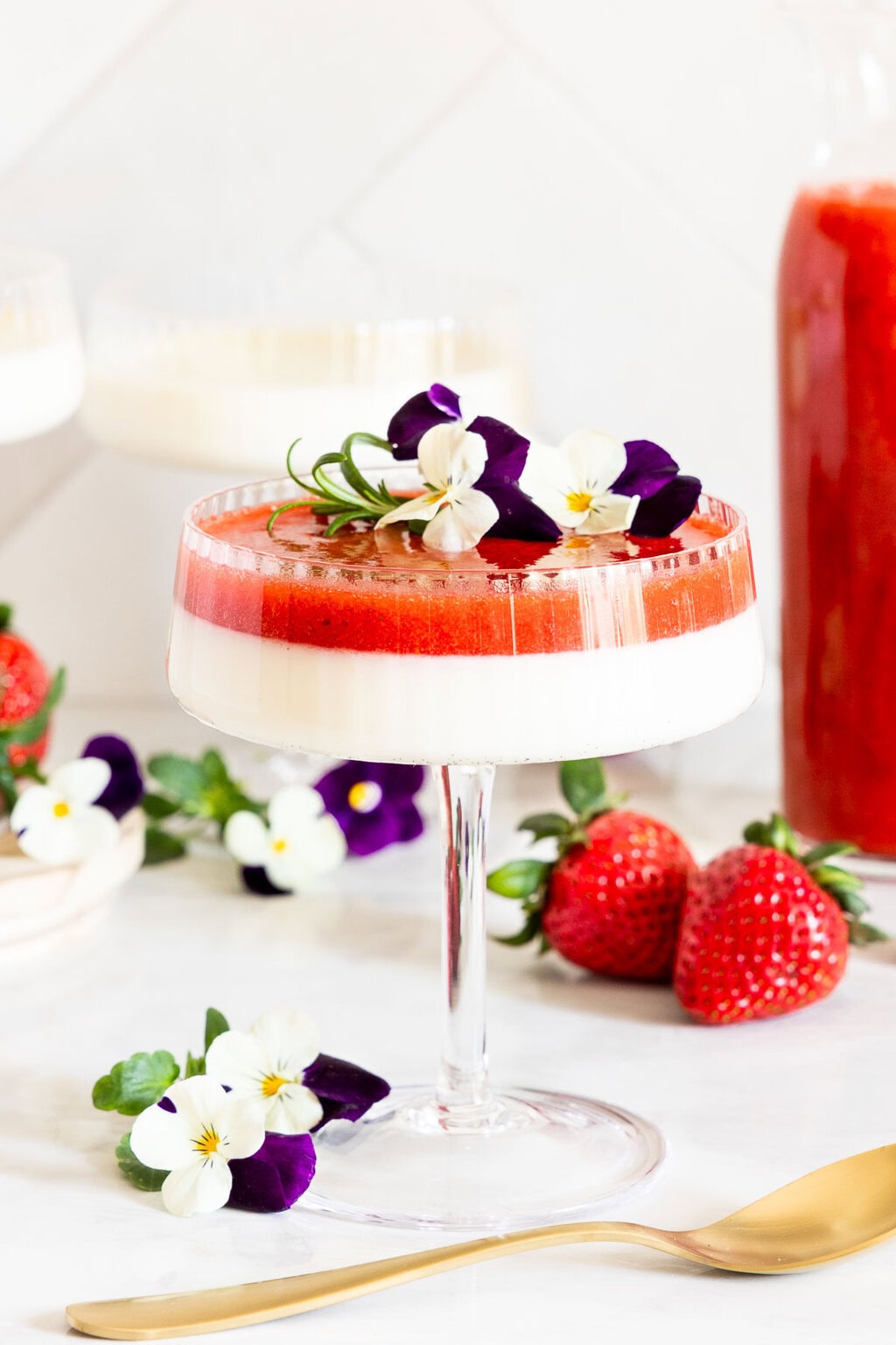 A long stemmed dessert glass of Ridiculously Easy Lemon Vanilla Bean Panna Cotta with Strawberry Coulis as the top layer garnished with edible pansies and a sprig of rosemary