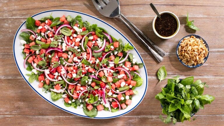 Horizontal overhead photo of an Asian Watermelon Herb Salad on a wood table with bowls of fresh mint and basil, slivered almonds and dressing.