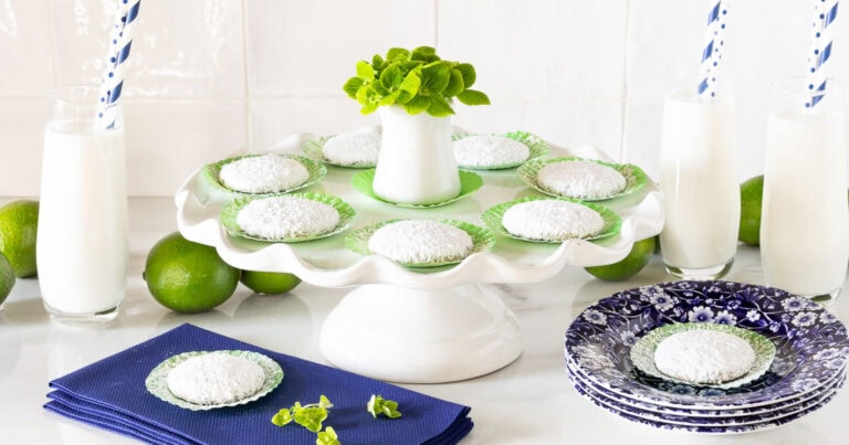 Horizontal photo of a batch of Key Lime Cooler Cookies on a white pedestal serving plate surrounded by glasses of milk.
