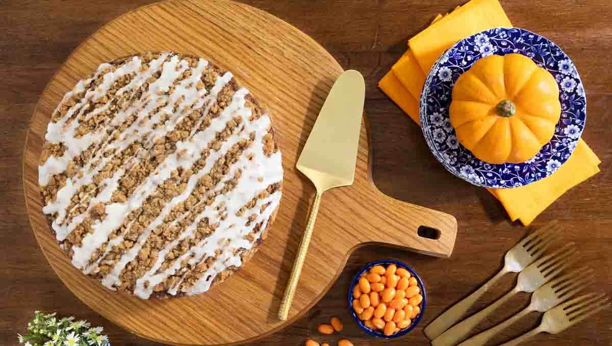 Bakery Style Pumpkin Crumb Cake - thecafesucrefarine.com Horizontal overhead photo of a Bakery Style Pumpkin Crumb Cake on a round wood serving board surrounded by pumpkin jelly beans and a mini pumpkin.