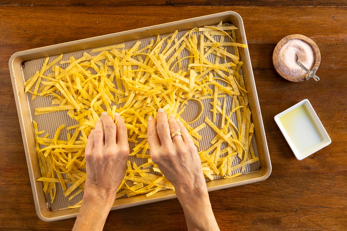 Overhead image of hands spreading thin strips of corn tortillas on a baking sheet, with olive oil and salt nearby, preparing homemade oven-baked tortilla strips.
