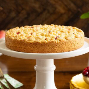 Horizontal photo of a Cranberry Pear Shortbread Tart on a white pedestal serving plate surrounded by fresh cranberries and pears.