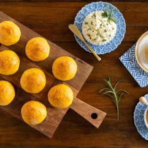 Horizontal overhead photo of a batch of No-Knead Sweet Potato Rolls on a wood cutting board featuring Herb Butter and honey.