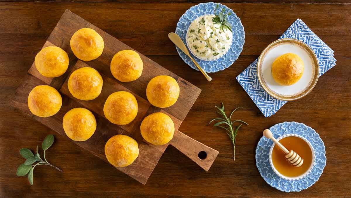 No-Knead Sweet Potato Rolls - thecafesucrefarine.com Horizontal overhead photo of a batch of No-Knead Sweet Potato Rolls on a wood cutting board featuring compound herb butter and honey.