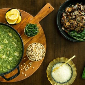 Horizontal overhead photo of a cast iron pot of Marry Me Chicken and Spinach Risotto with bowls of Parmesan cheese, caramelized mushrooms, lemon wedges and toasted pine nuts.