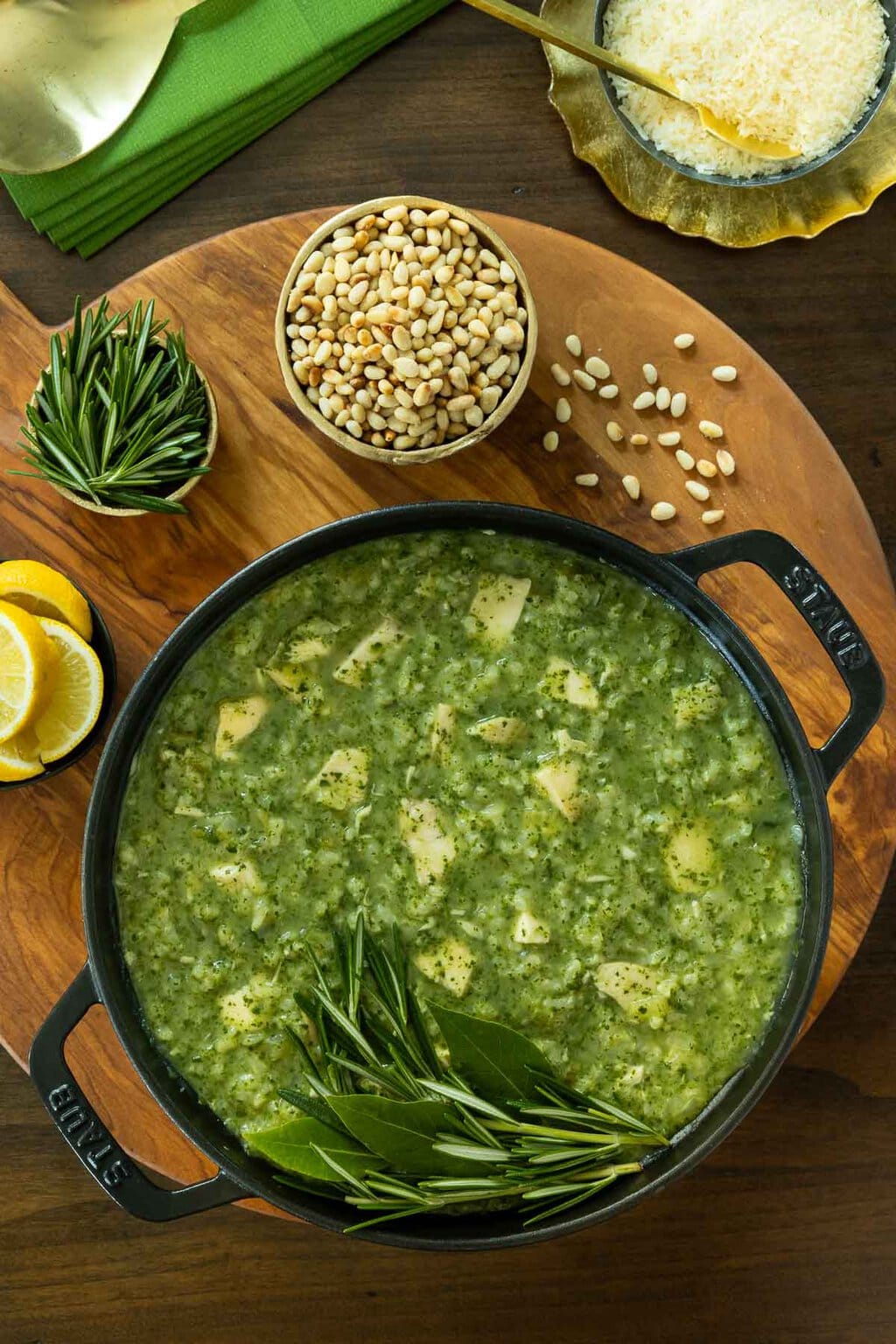 Vertical overhead photo of a cast iron pot of Marry Me Chicken and Spinach Risotto on a round wood plate surrounded by dishes of caramelized mushrooms, toasted pine nuts, Parmesan cheese, rosemary and lemon wedges.
