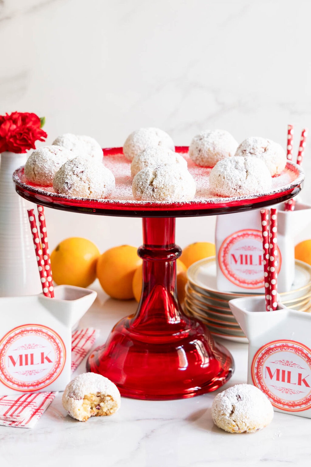 Orange Cranberry Amaretti Cookies on a red cake stand with oranges and cartons of milk.