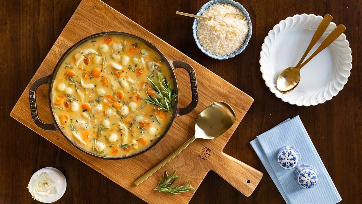 Horizontal overhead photo of a cast iron pot of Feel Better Chicken Gnocchi Soup on a wood board garnished with rosemary sprigs.