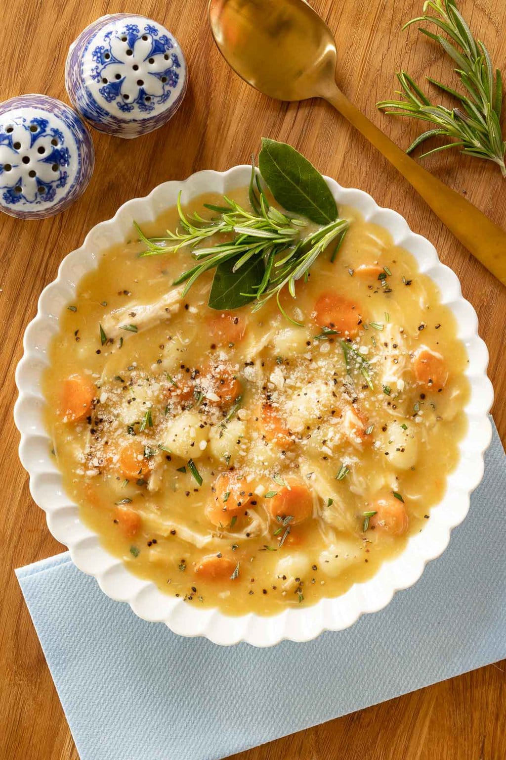 Overhead horizontal closeup photo of a bowl of Feel-Better Chicken Gnocchi Soup on a wood board garnished with sprinkles of cheese and rosemary sprigs.