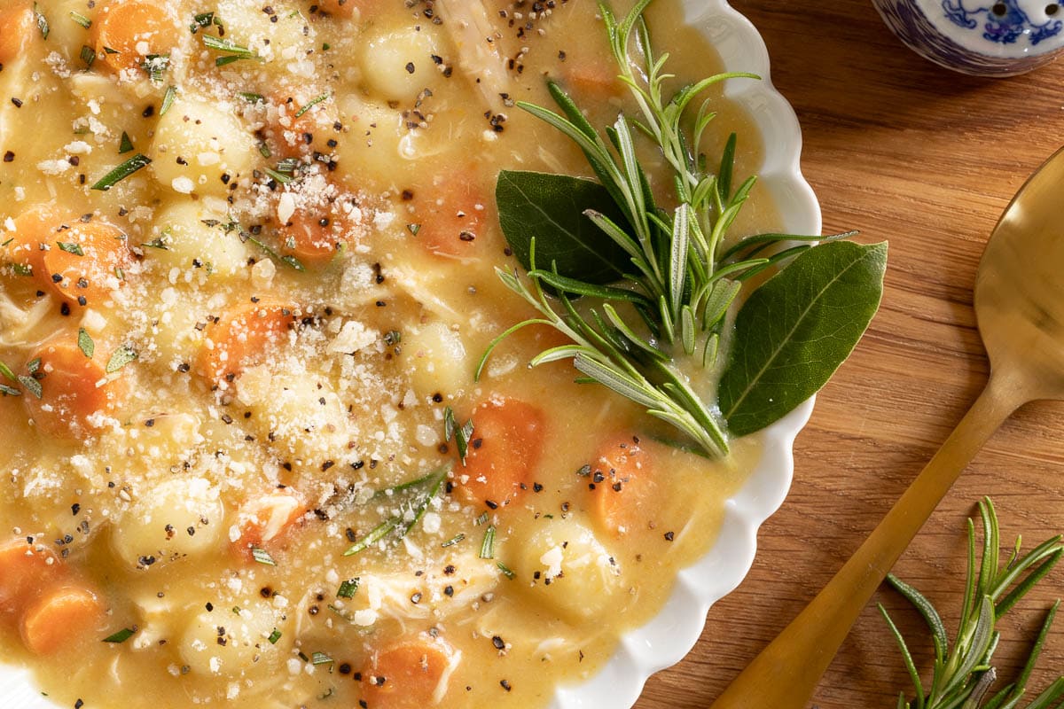 Horizontal extreme closeup photo of a bowl of Feel-Better Chicken Gnocchi Soup on a wood cutting board and garnished with sprigs of rosemary.