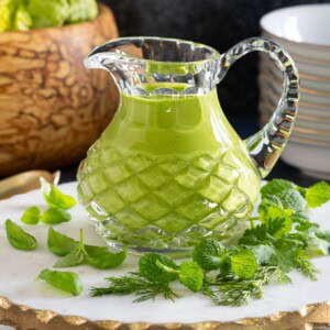 Horizontal photo of a glass cruet filled with Green Goddess Dressing and surrounded by fresh herb leaves with a bowl of salad in the background.