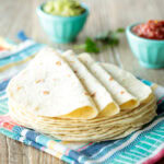 a pile of homemade flour tortillas on a colorful towel complete with little turquoise bowls of salsa and guacamole