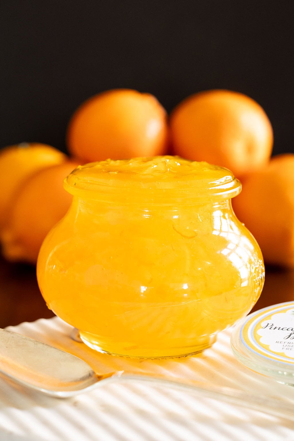 Pineapple Mandarin Marmalade - thecafesucrefarine.com Vertical extreme closeup photo of a Weck jar of Pineapple Mandarin Marmalade on a marble plate with Mandarin oranges in the background.