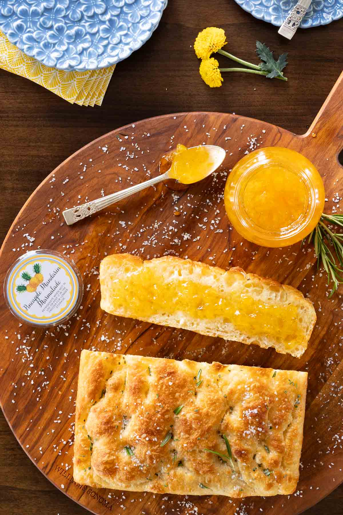 Pineapple Mandarin Marmalade - thecafesucrefarine.com Vertical overhead photo of a jar of Pineapple Mandarin Marmalade on a wood cutting board with a loaf of focaccia bread and a slice spread with the marmalade.