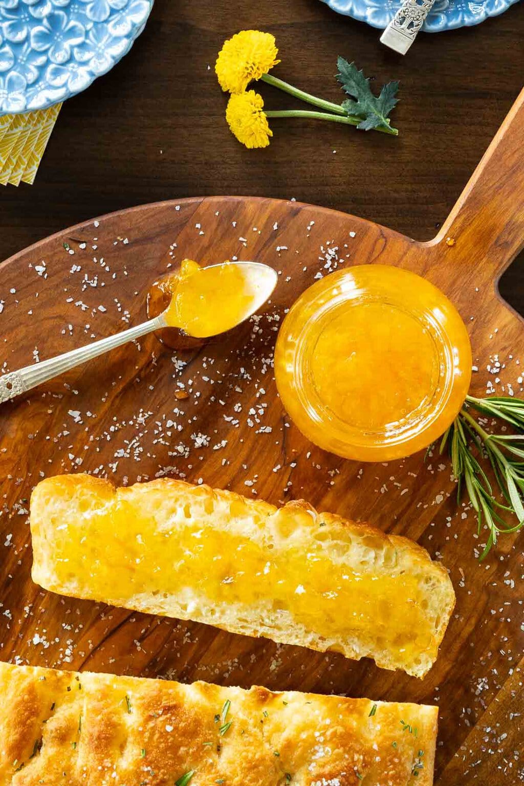 Pineapple Mandarin Marmalade - thecafesucrefarine.com Vertical overhead closeup photo of a jar of Pineapple Mandarin Marmalade on a round wood platter with a slice of focaccia bread spread with the marmalade.