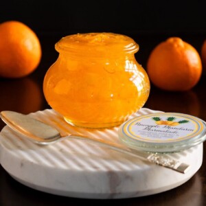 Horizontal closeup photo of a Weck jar of Pineapple Mandarin Marmalade on a round marble plate with Mandarin oranges in the background.