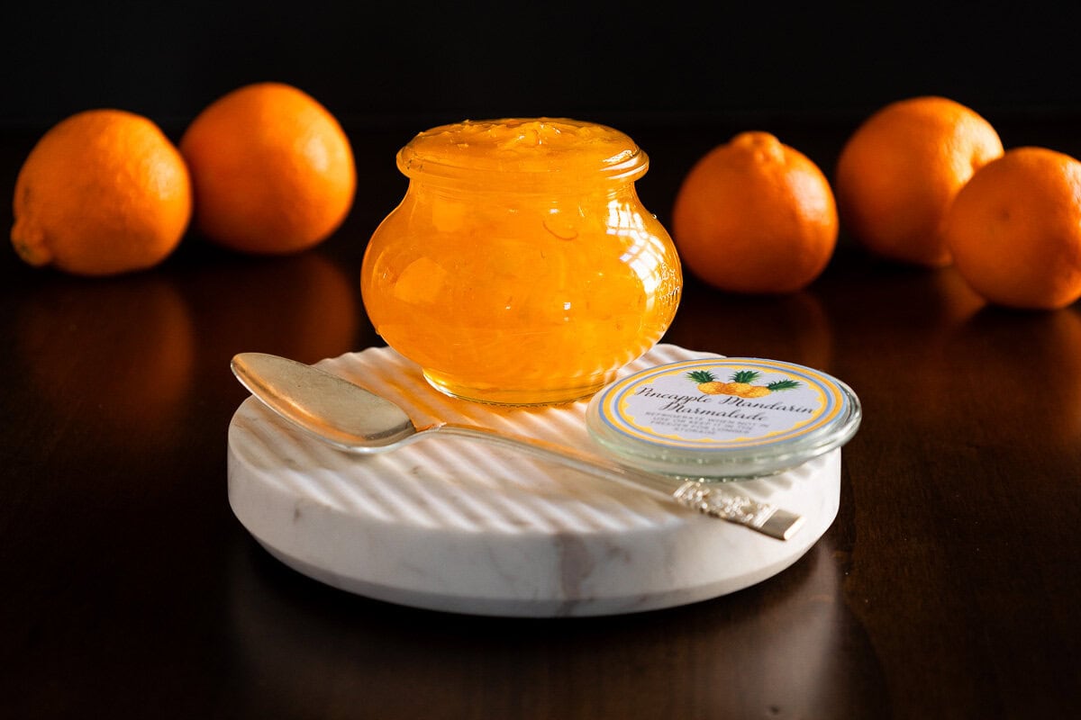 A horizontal closeup photo of a jar of Pineapple Mandarin Marmalade on a round marble platter.