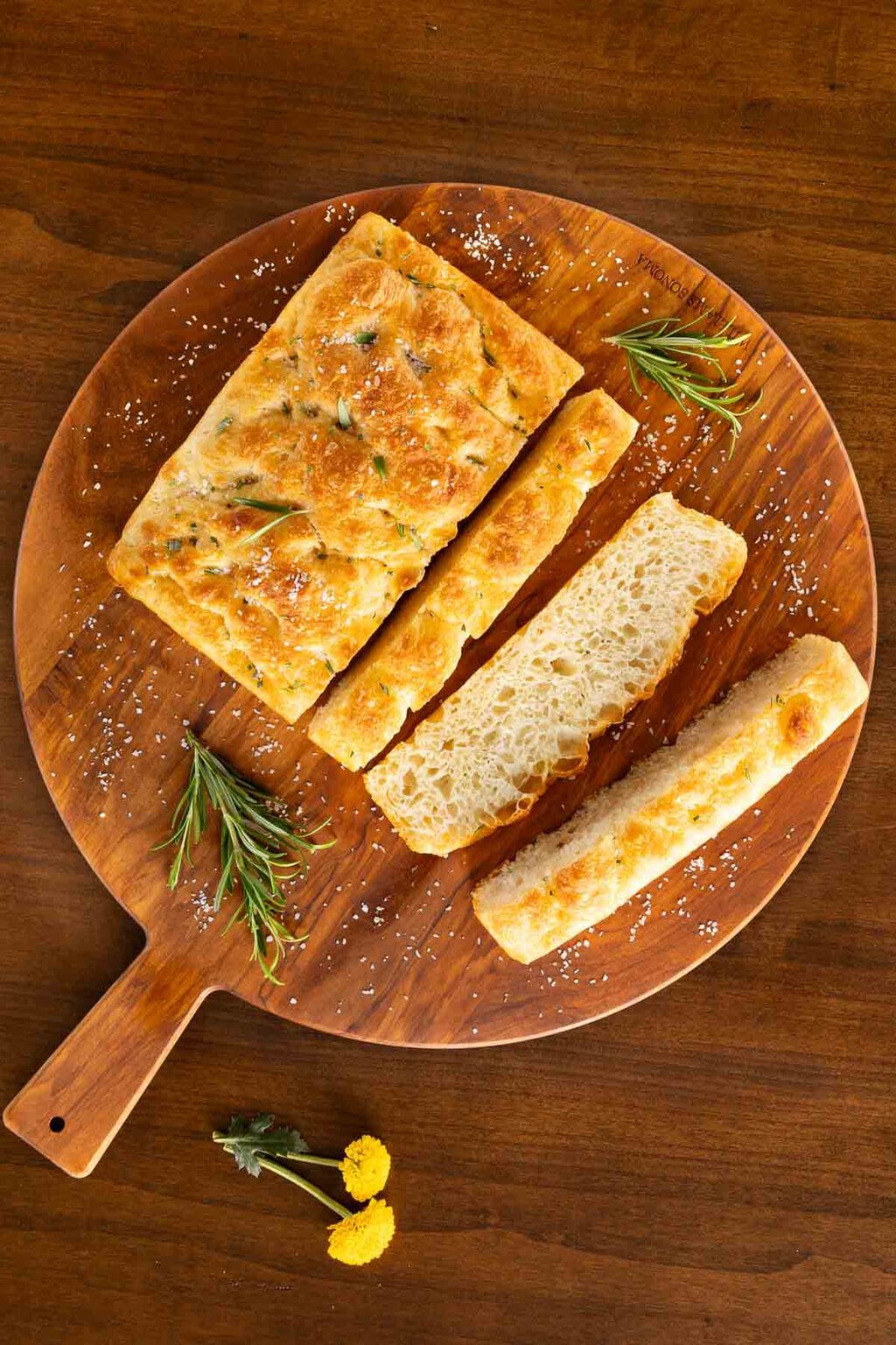Vertical overhead photo of a sliced loaf of Potato Focaccia (Focaccia Pugliese) on a round wood cutting board.