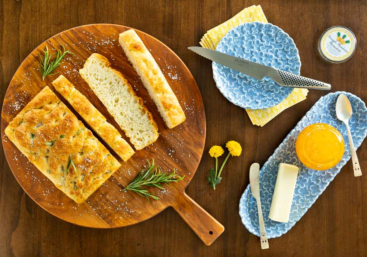 Horizontal overhead photo of a sliced loaf of Potato Focaccia (Focaccia Pugliese) on a round cutting board with a jar of Pineapple Mandarin Marmalade and a stick of butter.