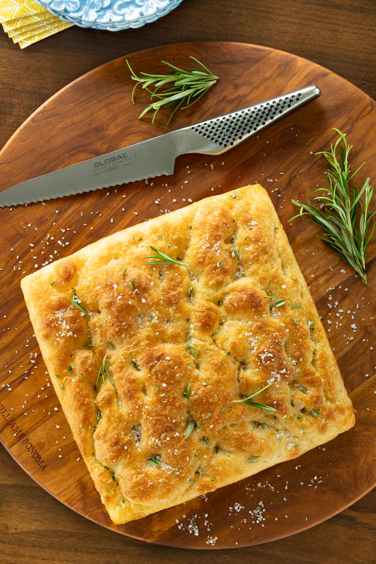 Vertical overhead photo of a loaf of Potato Focaccia (Focaccia Pugliese) on a round wood cutting board with sprigs of rosemary surrounding the bread.