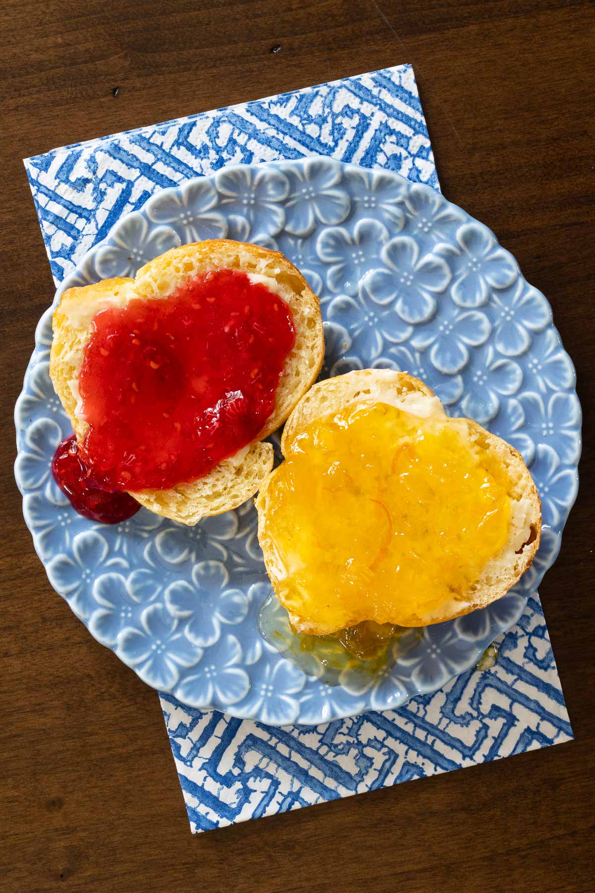 a closeup view of a no knead challah roll cut in half and spread with two different jams, one red and one yellow sitting on a blue flowered plate