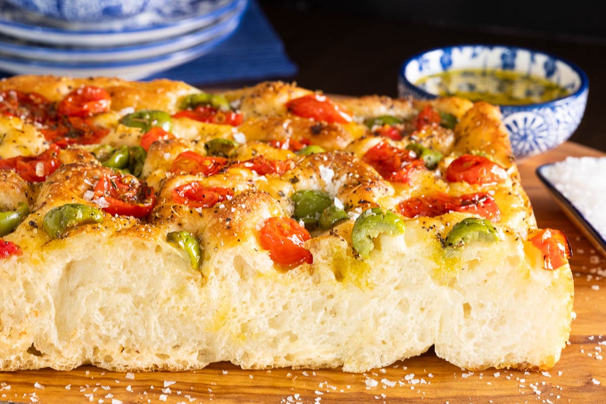 Ridiculously Easy Tomato Olive Focaccia - thecafesucrefarine.com Horizontal closeup photo of the side and top of a loaf of Ridiculously Easy Tomato Olive Focaccia bread on a wooden cutting board.