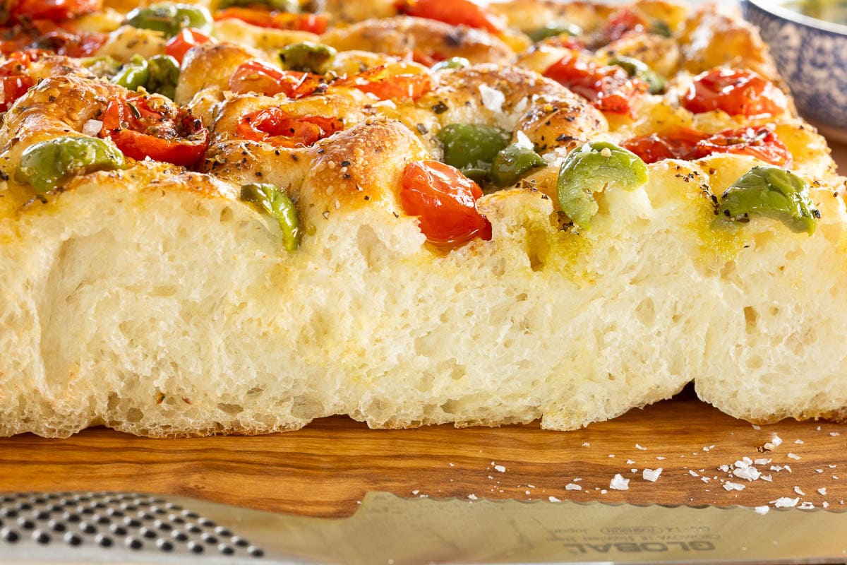 Ridiculously Easy Tomato Olive Focaccia - thecafesucrefarine.com Horizontal extreme closeup photo of the side of a slice of Ridiculously Easy Tomato Olive Focaccia on a wood cutting board.