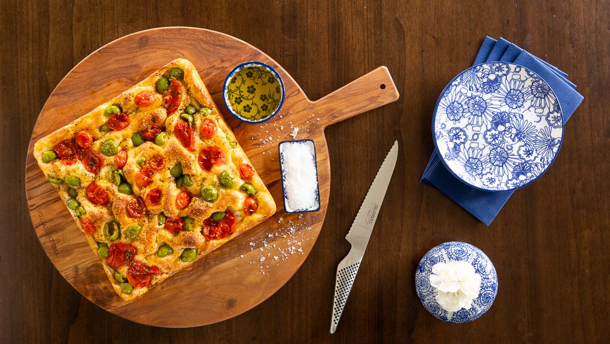 Ridiculously Easy Tomato Olive Focaccia - thecafesucrefarine.com Horizontal overhead photo of a loaf of Ridiculously Easy Tomato Olive Focaccia on a round wood cutting board.