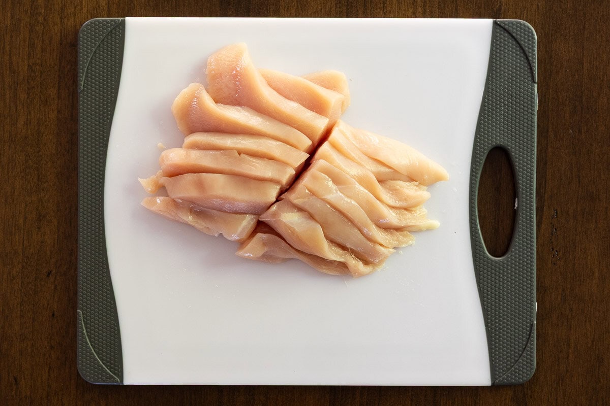 Overhead photo of a chicken breast cut into slices going across the grain and cut in half on a plastic cutting board.