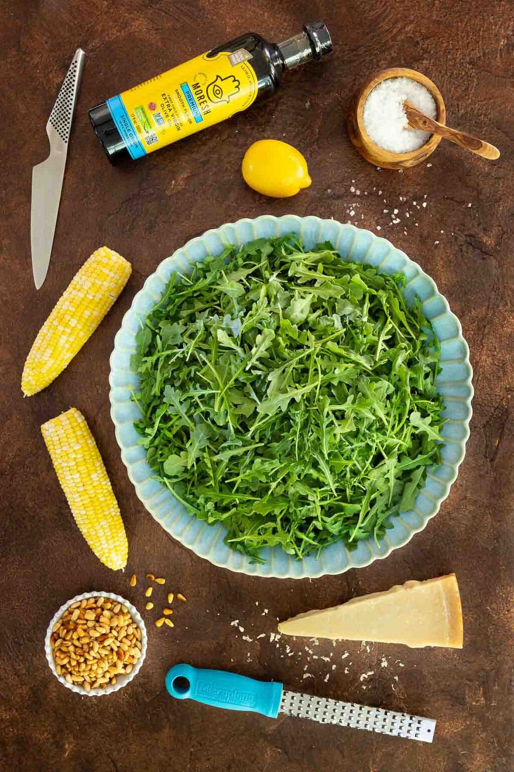 Flat lay of arugula in a blue bowl surrounded by salad ingredients including fresh corn, parmesan, pine nuts, olive oil, lemon, and sea salt