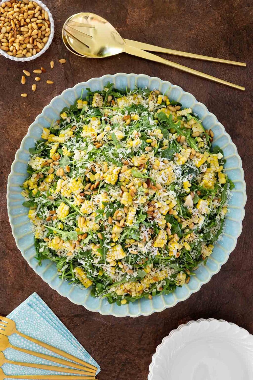 Arugula corn salad in a blue scalloped bowl topped with shaved parmesan and pine nuts, surrounded by gold serving utensils and salad plates