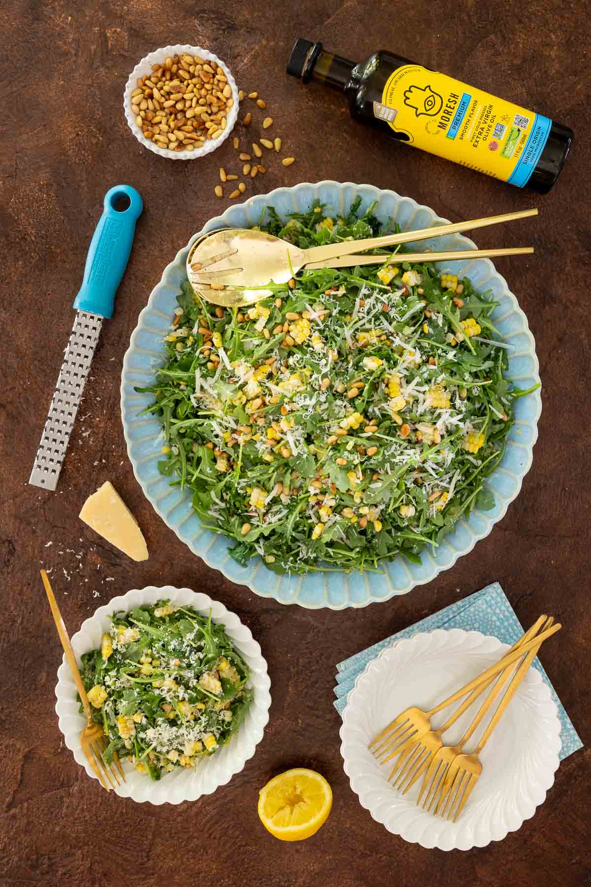 Overhead image of arugula corn salad in a scalloped bowl with parmesan, pine nuts, and lemon, surrounded by serving plates, olive oil, and fresh ingredients