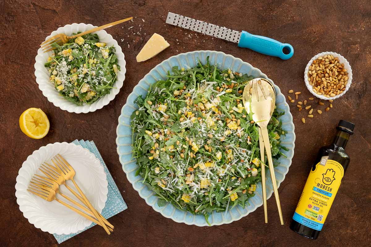 Overhead view of arugula corn salad topped with shaved parmesan and toasted pine nuts in a blue serving bowl, surrounded by salad plates, lemon, pine nuts, and a bottle of olive oil