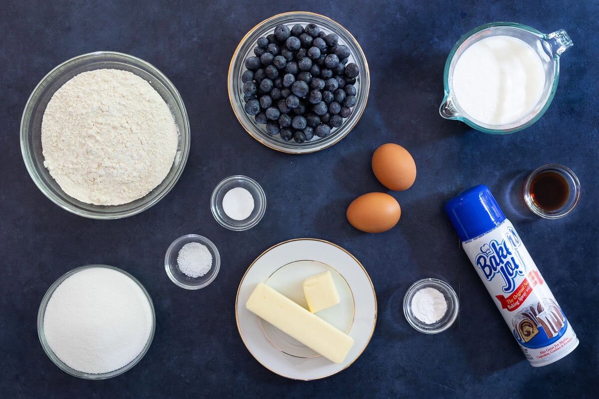 Overhead view of ingredients for this recipe arranged on a dark surface, including flour, blueberries, buttermilk, butter, eggs, sugar, vanilla, salt, baking powder, and baking spray