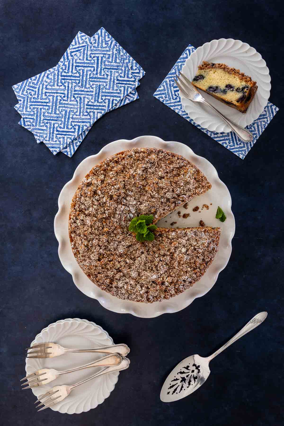 Overhead view of this coffee cake with one slice removed and plated, showing the moist interior and juicy blueberries, styled with dessert forks and napkins