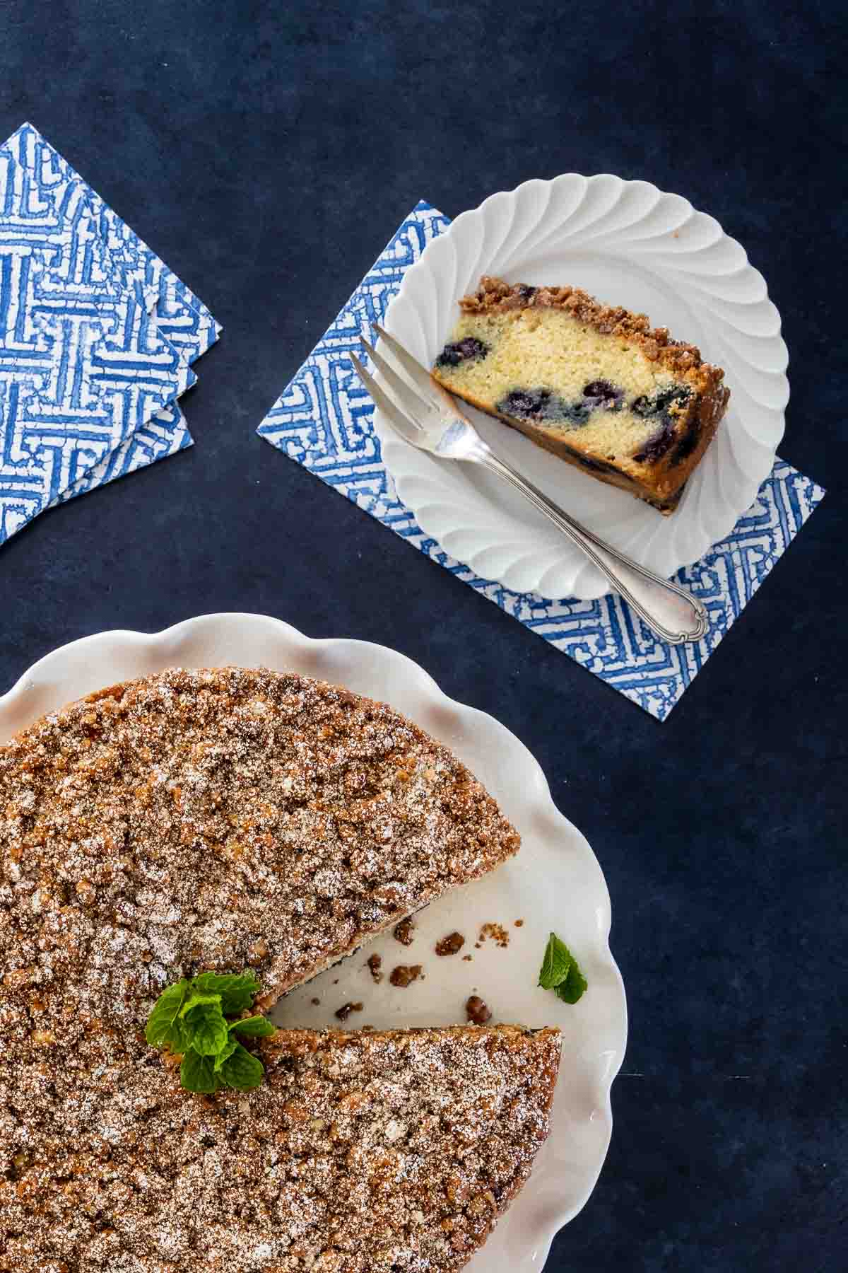 A slice of blueberry coffee cake on a white plate showing the moist interior and vibrant berries, next to the remaining streusel-topped cake with one slice removed
