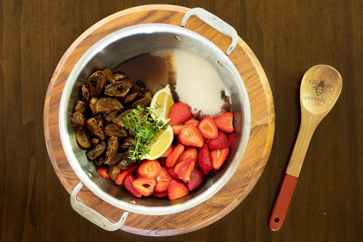 Stainless steel pot with ingredients for strawberry balsamic fig jam: figs, fresh strawberries, fresh thyme, balsamic vinegar, lemon wedges adn sugar.