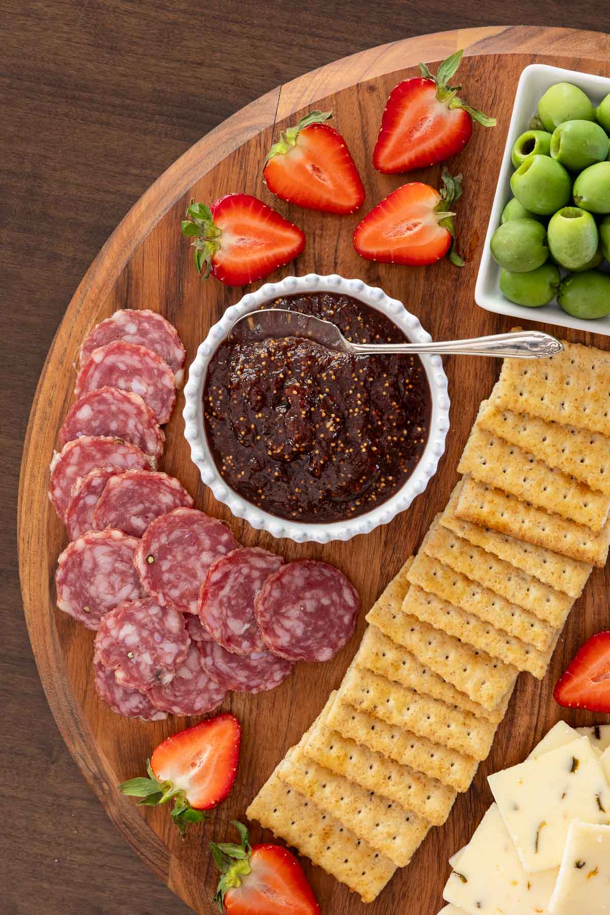 Close-up of of a charcuterie board with a small bowl of strawberry balsamic fig jam, crackers, sliced sopressata, olives, and fresh strawberries for garnish.