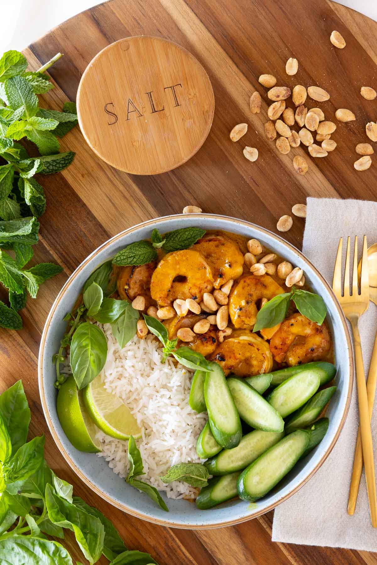 Bowl of Thai Coconut Curry Shrimp with basmati rice, sliced cucumbers, roasted peanuts, fresh mint and basil, served on a wooden board with gold flatware.