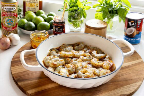 Raw shrimp tossed with spices in a white braiser pan, surrounded by ingredients for Thai coconut curry sauce including red curry paste, coconut milk, limes, garlic, shallots, and fresh herbs.