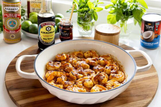 Broiled shrimp in a white braiser pan, surrounded by Thai coconut curry sauce ingredients including soy sauce, red curry paste, coconut milk, olive oil, honey, and fresh herbs.