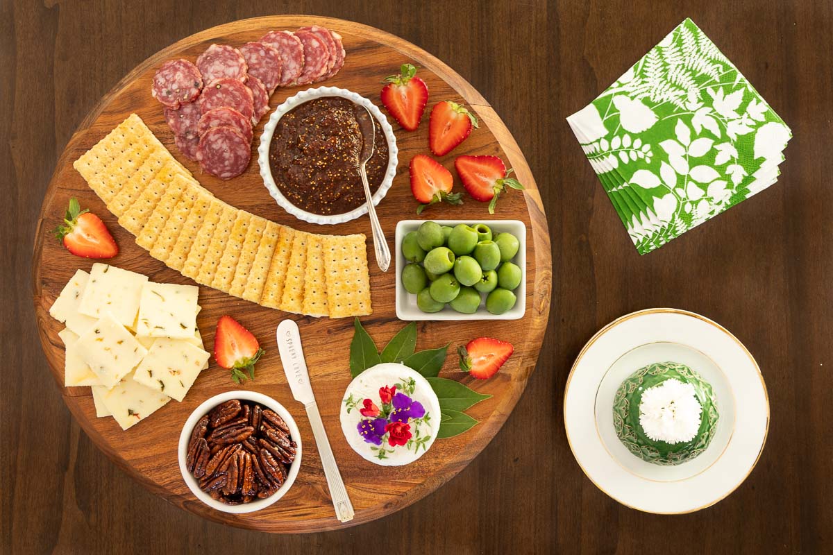 Charcuterie board with a small bowl of strawberry balsamic fig jam, crackers, sliced sopressata, green olives, goat cheese, candied pecans, sliced cheese, and fresh strawberries for garnish.