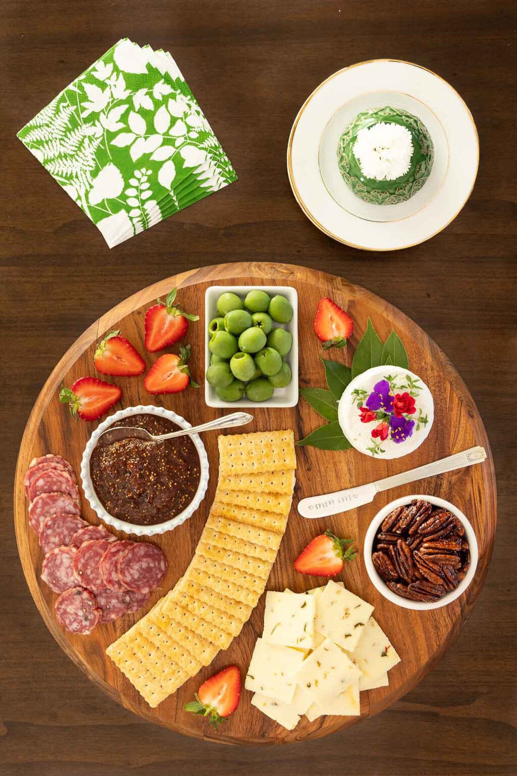 Charcuterie board with a small bowl of strawberry balsamic fig jam, crackers, sliced sopressata, green olives, goat cheese, candied pecans, sliced cheese, and fresh strawberries for garnish.